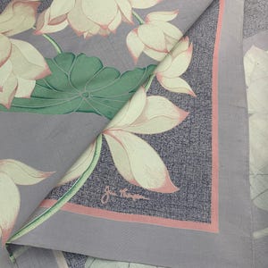 May include: A folded, square textile featuring a floral pattern of lotus blossoms. The flowers are cream-colored with pink edges, set against a gray background with a pink border. The textile has a signature in the corner.