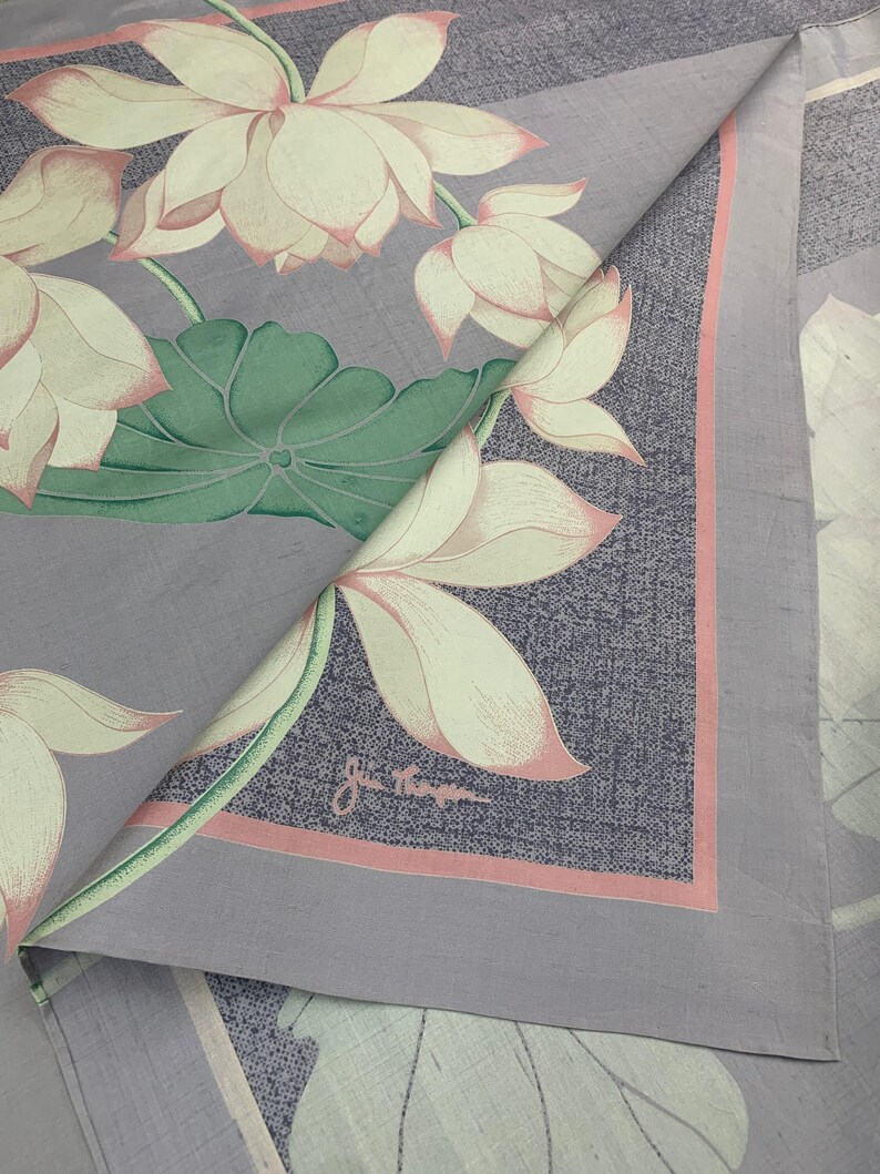 May include: A folded, square textile featuring a floral pattern of lotus blossoms. The flowers are cream-colored with pink edges, set against a gray background with a pink border. The textile has a signature in the corner.