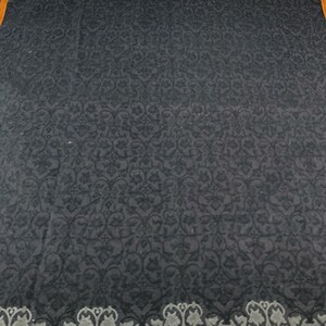 Puede incluir: Un textil gris oscuro con un patr&oacute;n floral repetitivo. El borde presenta un dise&ntilde;o decorativo en gris y blanco, con un ribete naranja. El textil es cuadrado y parece ser un art&iacute;culo decorativo.