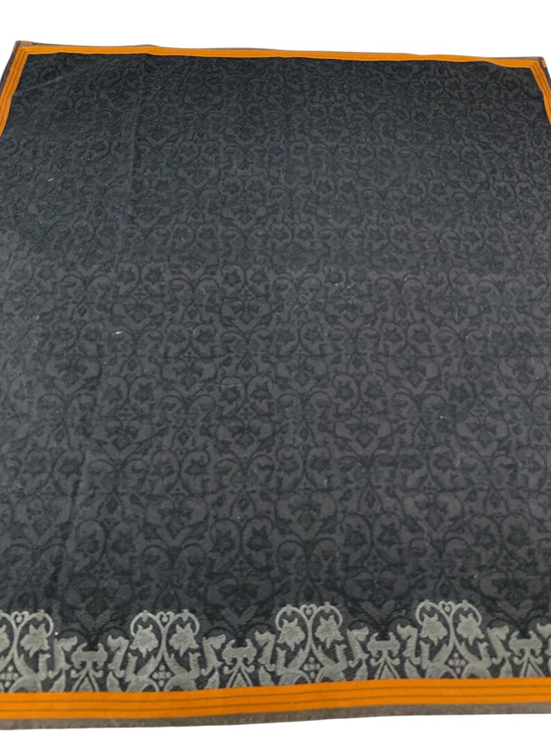 Puede incluir: Un textil gris oscuro con un patr&oacute;n floral repetitivo. El borde presenta un dise&ntilde;o decorativo en gris y blanco, con un ribete naranja. El textil es cuadrado y parece ser un art&iacute;culo decorativo.