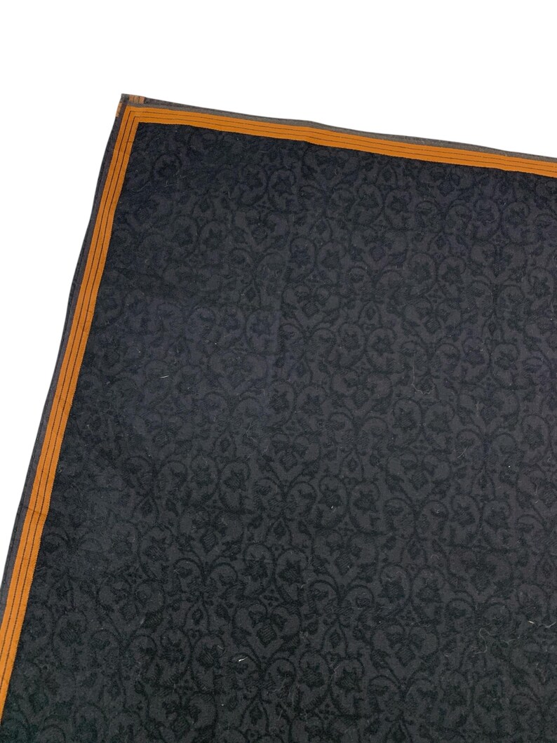 Puede incluir: Una secci&oacute;n de un textil gris oscuro con un patr&oacute;n floral repetitivo. El borde est&aacute; bordeado con una fina franja naranja. El textil tiene un brillo sutil, lo que sugiere una tela lujosa.