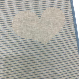 K&ouml;nnte beinhalten: Ein quadratischer, hellfarbener Stoff mit blauem Rand und blauen horizontalen Streifen. In der Mitte ist eine gro&szlig;e Herzform zu sehen. Der Stoff scheint Leinen oder ein &auml;hnliches Material zu sein.