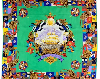 Puede incluir: Pañuelo cuadrado de seda con temática náutica. El pañuelo presenta un diseño central de un velero, rodeado por un borde decorativo con brújulas, banderas y detalles de cuerda. La paleta de colores incluye verde, dorado, azul, rojo y morado.