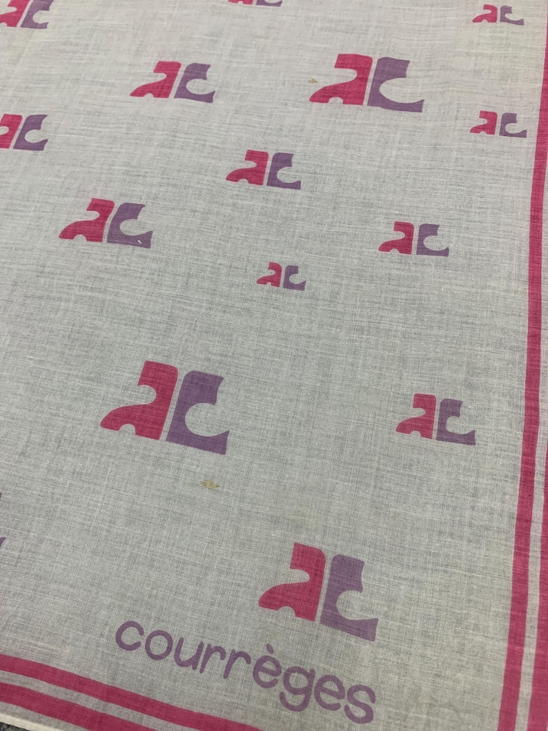 Peut inclure: Un mouchoir Courr&egrave;ges gris clair avec un motif r&eacute;p&eacute;t&eacute; de logos "AC" roses et violets. La bordure et le texte "courr&egrave;ges" sont roses. Le tissu a une texture subtile.