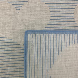 K&ouml;nnte beinhalten: Nahaufnahme eines hellgrauen Stoffes mit einem sich wiederholenden Muster aus blauen horizontalen Linien und geometrischen Formen. Der Stoff hat ein weiches, strukturiertes Aussehen, mit einem blauen Rand.