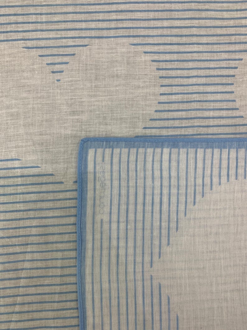 K&ouml;nnte beinhalten: Nahaufnahme eines hellgrauen Stoffes mit einem sich wiederholenden Muster aus blauen horizontalen Linien und geometrischen Formen. Der Stoff hat ein weiches, strukturiertes Aussehen, mit einem blauen Rand.