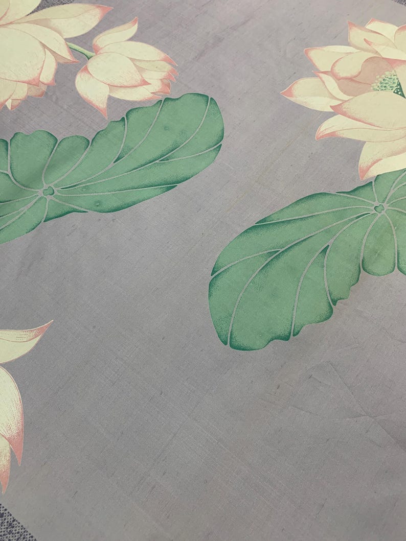 May include: A close-up of a fabric with a floral pattern. The design features large, light yellow lotus flowers with pink edges and green leaves on a gray background. The fabric has a subtle texture.