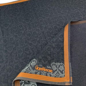 Puede incluir: Un pa&ntilde;uelo negro doblado con un estampado floral y un borde naranja. La esquina presenta el logotipo "Gianni Versace" en naranja y gris.