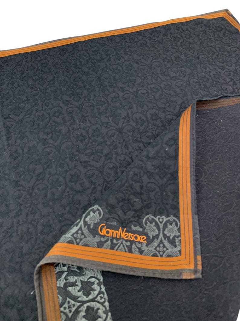 Puede incluir: Un pa&ntilde;uelo negro doblado con un estampado floral y un borde naranja. La esquina presenta el logotipo "Gianni Versace" en naranja y gris.