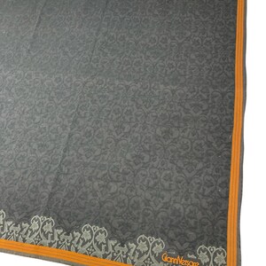 Puede incluir: Pa&ntilde;uelo cuadrado gris oscuro con un intrincado estampado floral. El pa&ntilde;uelo tiene un borde naranja y un dise&ntilde;o floral decorativo a lo largo de un borde. El logotipo de Gianni Versace es visible en la esquina.