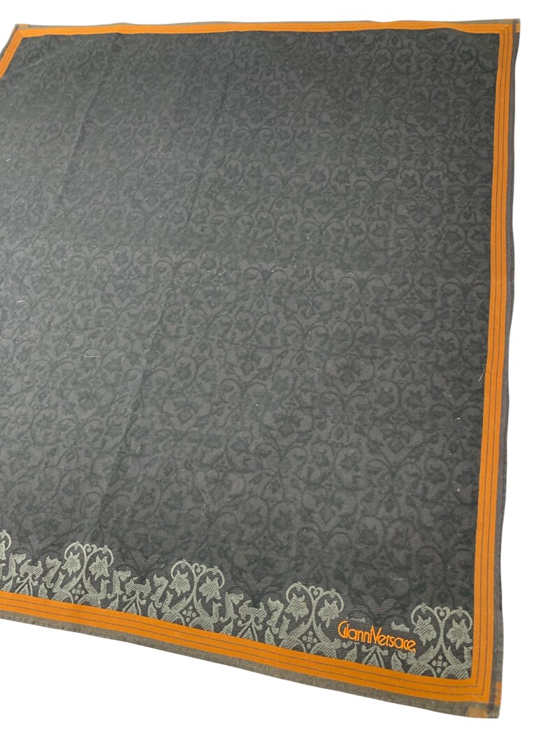 Puede incluir: Pa&ntilde;uelo cuadrado gris oscuro con un intrincado estampado floral. El pa&ntilde;uelo tiene un borde naranja y un dise&ntilde;o floral decorativo a lo largo de un borde. El logotipo de Gianni Versace es visible en la esquina.