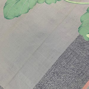 May include: A light gray textile with a botanical design. The image features large green leaves with white outlines and a gray border with a dotted pattern. A thin pink stripe accents the border.