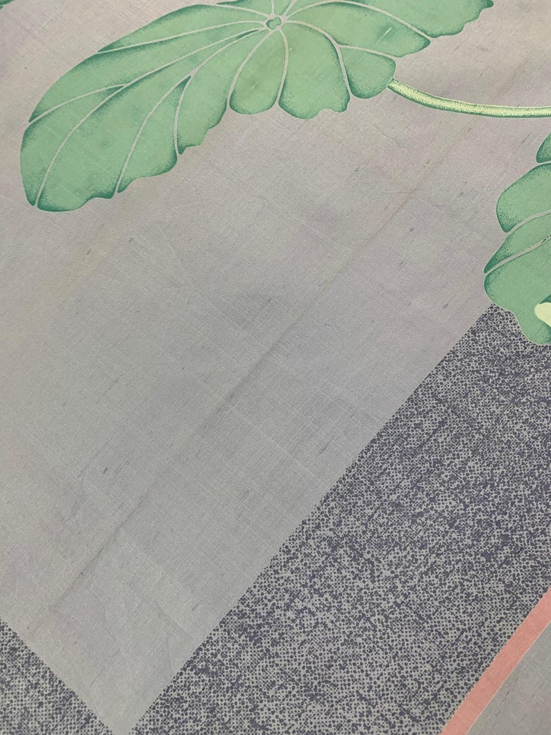 May include: A light gray textile with a botanical design. The image features large green leaves with white outlines and a gray border with a dotted pattern. A thin pink stripe accents the border.