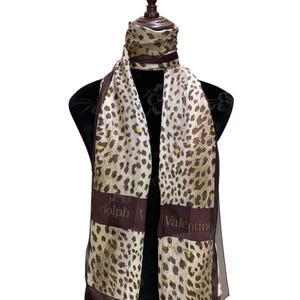 May include: A rectangular scarf featuring a beige and brown leopard print pattern with dark brown borders. The text Rudolph Valentino is printed in a light font across the lower dark brown sections of the scarf, which is draped around a mannequin.