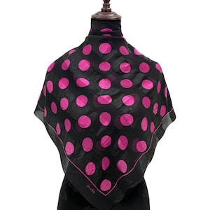 May include: Black square scarf with large pink polka dots. The scarf is draped over a mannequin. The scarf has a sheer appearance and a dark border. The text "jan etshila" is printed on the scarf.