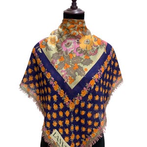 May include: A square Lanvin Paris scarf featuring a dark blue background with a repeating pattern of small orange flowers. The center displays a large floral motif in shades of pink, orange, and green. The scarf has frayed edges and a white label.
