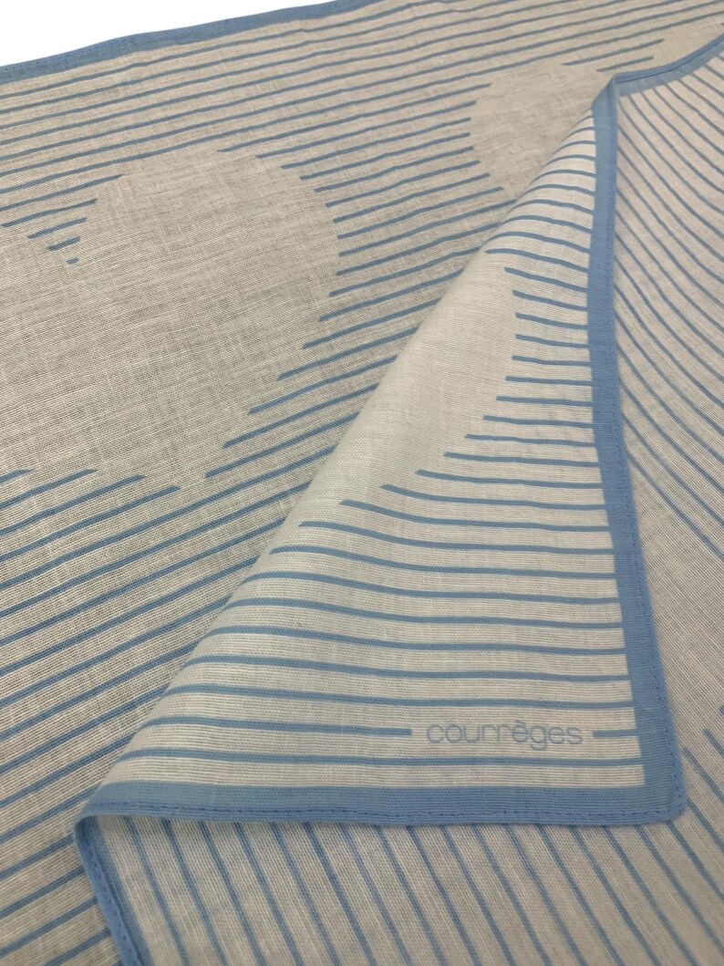 K&ouml;nnte beinhalten: Ein gefalteter hellgrauer Schal mit blauem Streifenmuster und blauem Rand. Das Wort "courr&egrave;ges" ist in kleinen, grauen Buchstaben in der unteren rechten Ecke des Schals aufgedruckt.