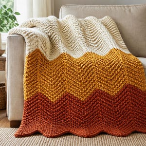Pode incluir: Uma manta de malha grossa com um padrão de chevron e secções horizontais de cores creme, amarelo mostarda e laranja queimado. A textura do fio grosso cria um design em ziguezague distinto em toda a superfície da manta.
