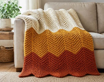 May include: A chunky knit throw blanket featuring a chevron pattern with horizontal colour-blocked sections in cream, mustard yellow, and burnt orange. The thick yarn texture creates a distinct zigzag design across the entire surface of the blanket.