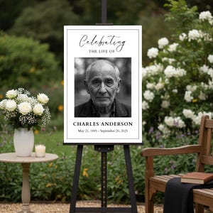 May include: A black easel displays a memorial sign with the text "Celebrating the Life of Charles Anderson." The sign features a black and white portrait of an older man and his birth and death dates. White flowers and a candle sit on a small table.