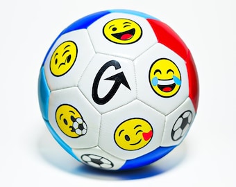 Könnte beinhalten: Ein weißer Fußball mit verschiedenen gelben Emoji-Gesichtern und schwarzen Fußballgrafiken. Der Ball hat blaue und rote Paneele. Ein großes schwarzes "G"-Logo ist vorhanden.