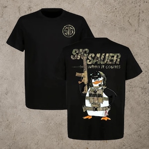 Könnte beinhalten: Ein schwarzes Kurzarm-T-Shirt mit einem kleinen Sig Sauer-Logo auf der Brust. Die Rückseite zeigt eine große Grafik eines Pinguins in taktischer Ausrüstung mit einem Gewehr sowie den Text Sig Sauer und den Schriftzug when it counts.