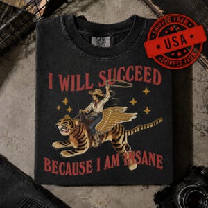 May include: A charcoal grey crewneck t-shirt featuring a graphic of a person in a cowboy hat riding a winged tiger while holding a lasso. The text reads I WILL SUCCEED BECAUSE I AM INSANE in red lettering, accented by yellow stars on the dark fabric.