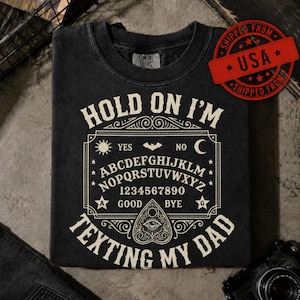 May include: A black short-sleeve t-shirt featuring a cream-coloured graphic of a spirit board with the alphabet, numbers, and symbols. The text reads "HOLD ON I'M TEXTING MY DAD" in a bold, arched font surrounding the central spirit board design.