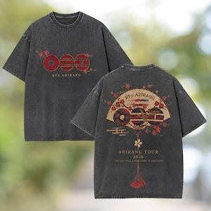 Op de afbeelding: Een antracietgrijs acid-wash T-shirt met een vooropdruk van rode cirkelvormige symbolen en de tekst BTS ARIRANG. De achterkant toont een grote illustratie van een waaier met rode bloemen, de tekst BTS ARIRANG, ARIRANG TOUR 2026 en namen.