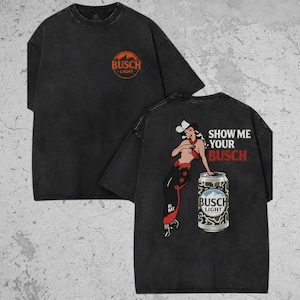 May include: A black short-sleeve t-shirt featuring an orange Busch Light logo on the front chest. The back displays a graphic of a cowgirl leaning against a Busch Light beer can with the text SHOW ME YOUR BUSCH in white and red lettering.