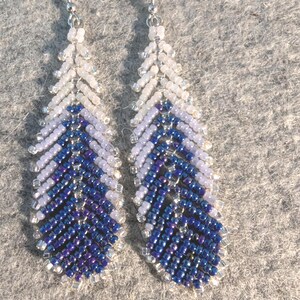 May include: A pair of beaded earrings in a feather design. The earrings feature a gradient of colors, transitioning from white at the top to light purple, and finally to a deep blue at the bottom. The earrings are suspended from silver-colored hooks.