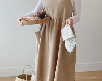 May include: A beige apron with a square neckline and large pockets. The apron is worn over a white long-sleeved top and white trousers. A woven basket and a checkered tea towel are also visible.