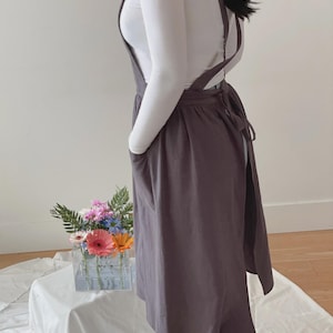 May include: A dark gray apron with cross-back straps and large front pockets. The apron is tied at the waist and worn over a white long-sleeved shirt. The apron is knee-length and made of a soft fabric.