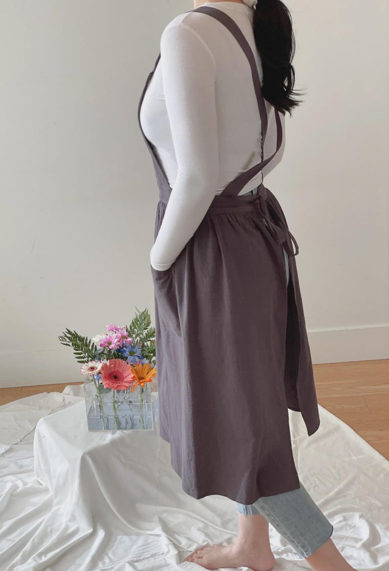 May include: A dark gray apron with cross-back straps and large front pockets. The apron is tied at the waist and worn over a white long-sleeved shirt. The apron is knee-length and made of a soft fabric.