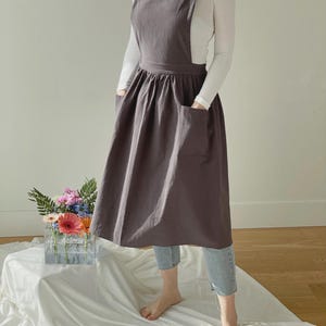 May include: A charcoal gray apron with a square neckline and large front pockets. The apron has a full skirt and suspender straps. The person is wearing a white long-sleeve shirt and light blue jeans. A bouquet of flowers sits in a clear vase.