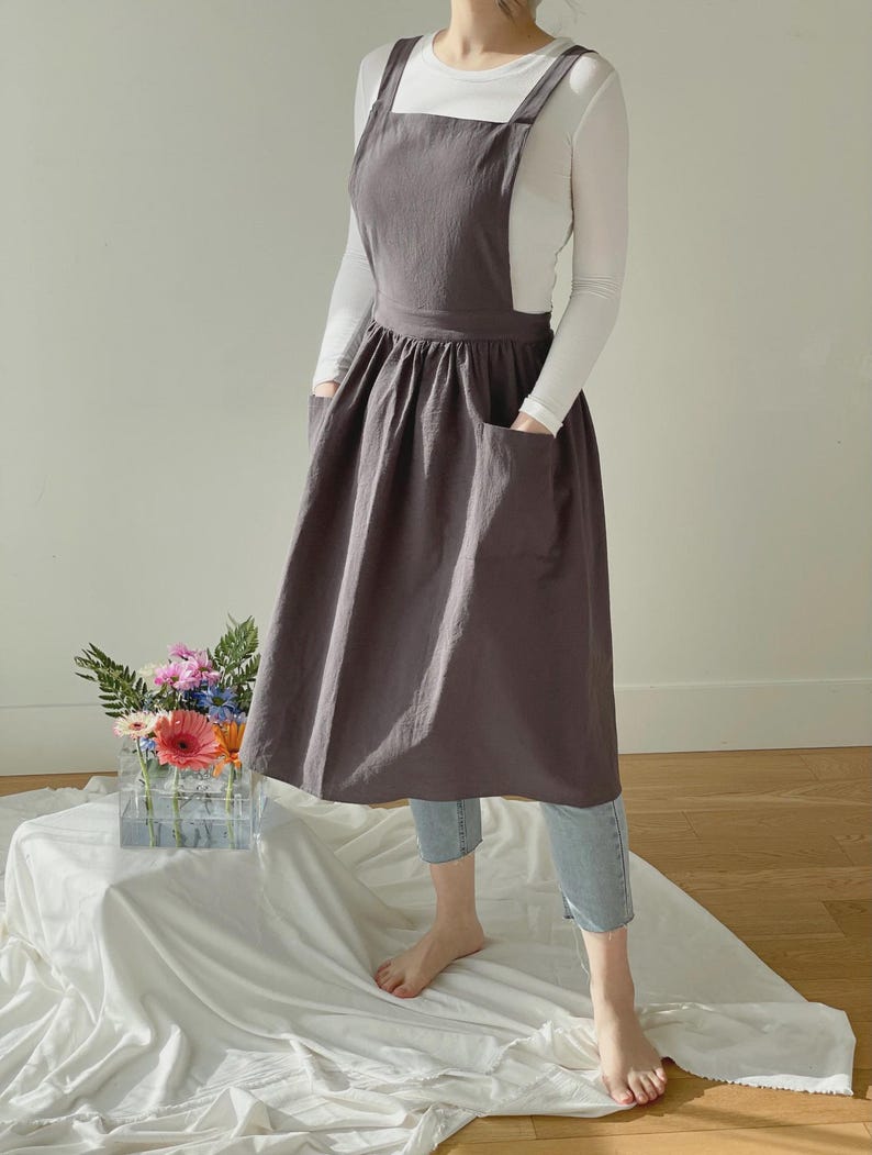 May include: A charcoal gray apron with a square neckline and large front pockets. The apron has a full skirt and suspender straps. The person is wearing a white long-sleeve shirt and light blue jeans. A bouquet of flowers sits in a clear vase.