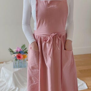 May include: A dusty rose apron with a bib, two large front pockets, and a waist tie. The apron has wide shoulder straps and is worn over a white long-sleeved shirt. The apron is made of a soft, textured fabric.