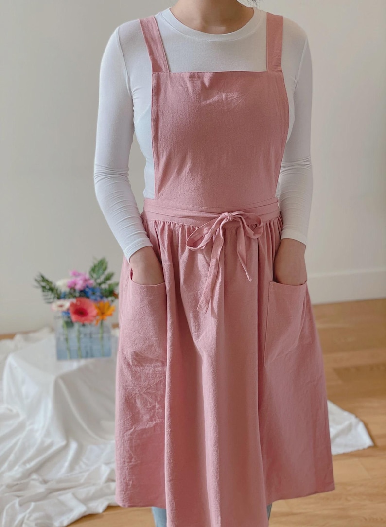 May include: A dusty rose apron with a bib, two large front pockets, and a waist tie. The apron has wide shoulder straps and is worn over a white long-sleeved shirt. The apron is made of a soft, textured fabric.