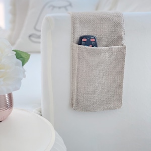 May include: A beige burlap bedside caddy with a pocket, hanging on a white headboard. A black remote control is in the pocket. A white flower and a small white table are in the foreground.