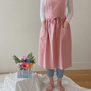 May include: A light pink apron with a cross-back design and two large front pockets. The apron is tied at the waist with a matching fabric tie. The model is wearing a white long-sleeve shirt and blue jeans.