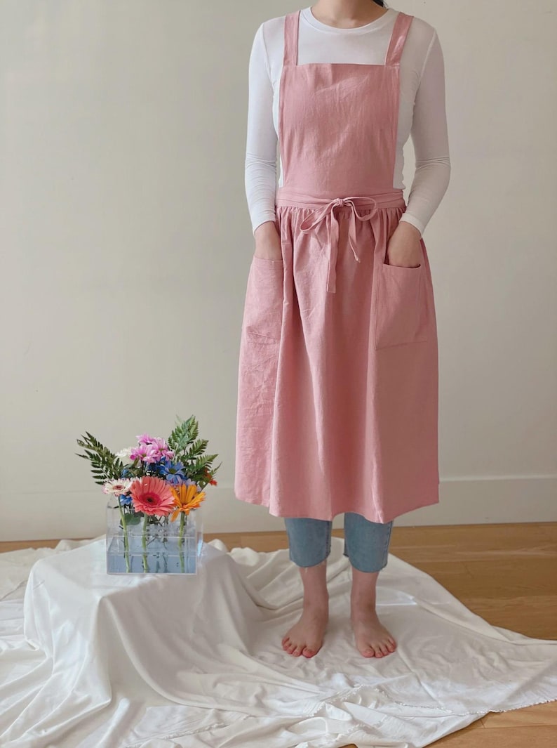 May include: A light pink apron with a cross-back design and two large front pockets. The apron is tied at the waist with a matching fabric tie. The model is wearing a white long-sleeve shirt and blue jeans.