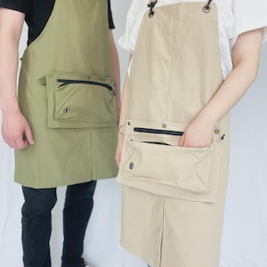 May include: Two aprons are shown, one olive green and one beige. Each apron features a large front pocket with a black zip and adjustable straps. The aprons are worn by two people, one in a black top and the other in a white top.