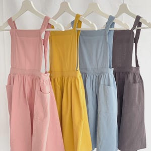 May include: Four aprons in pastel colors hang on a white rack. The aprons are in shades of pink, yellow, blue, and gray. Each apron has a large front pocket and long ties. The aprons are made of a soft fabric.