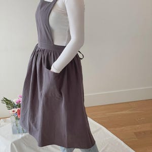 May include: A dark gray apron dress with a large front pocket and cross-back straps. The apron is worn over a white long-sleeved shirt and blue jeans. The apron is made of a soft, flowing fabric.