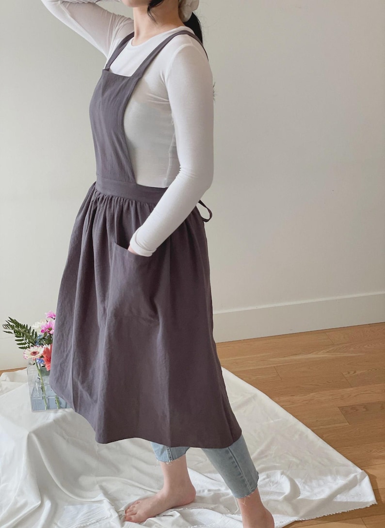 May include: A dark gray apron dress with a large front pocket and cross-back straps. The apron is worn over a white long-sleeved shirt and blue jeans. The apron is made of a soft, flowing fabric.