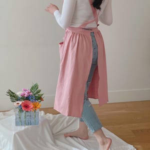 May include: A light pink apron with a cross-back design and two front pockets. The apron is long and covers the legs. The person is wearing a white long-sleeve shirt and blue jeans. A clear vase with colorful flowers sits on a white cloth.