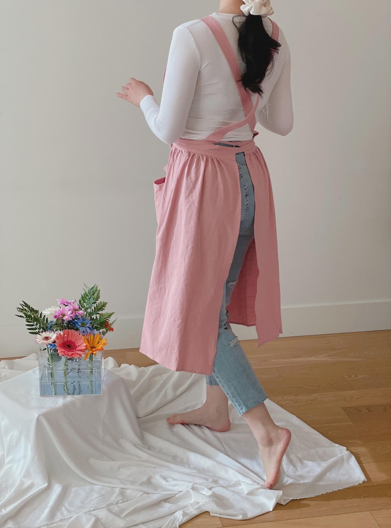 May include: A light pink apron with a cross-back design and two front pockets. The apron is long and covers the legs. The person is wearing a white long-sleeve shirt and blue jeans. A clear vase with colorful flowers sits on a white cloth.