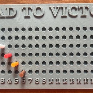 May include: A grey board game with the text "ROAD TO VICTORY" at the top. The board has a grid of holes with coloured pegs inserted. Numbers 1-20 are printed at the bottom. Peg colours include green, blue, white, red, orange, and brown.