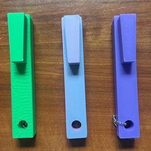 May include: Five colorful, rectangular, 3D-printed pen holders in pink, green, light blue, purple, and blue. Each has a rectangular clip at the top and a hole at the bottom. The pen holders are arranged in a row on a wooden surface.