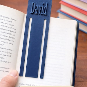 以下が含まれることがあります： 太字で「David」という名前が入った、ネイビーブルーのパーソナライズされたブックマーク。ブックマークは本に挟まれており、「PageKeeper」と「Personalized Name Bookmarks」の文字が見えます。